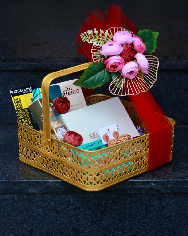 Gift hamper in a gold basket with red tulle, pink flowers and beauty gifts on dark stone